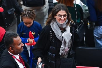 Fayza Lamari portant un manteau noir et un foulard gris à l'aéroport.
