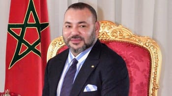 Roi Mohammed VI assis sur un fauteuil doré avec drapeau marocain en arrière-plan.