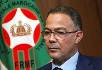 Fouzi Lekjaa en costume avec le logo du Maroc en arrière-plan.