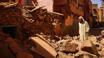Homme en vêtements traditionnels devant débris de bâtiment effondré.