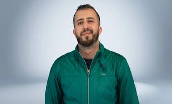 Anes Tina souriant, portant une veste verte, dans un studio neutre.