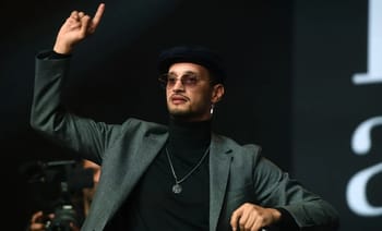 Soolking en costume avec lunettes, levant le doigt lors d'une intervention.