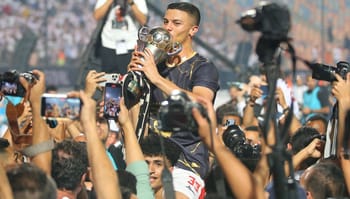 Joueur levant le trophée de la Coupe de la CAF entouré de fans et médias.