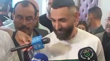 Karim Benzema souriant lors d'une visite en Algérie, entouré de médias et de personnes.