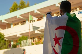 Homme tenant un drapeau algérien devant un bâtiment moderne.