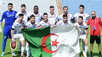 Groupe de joueurs de l'USM Alger avec drapeau algérien en fond.