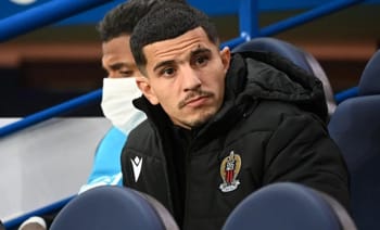 Youcef Atal assis sur le banc, portant un manteau noir avec le logo de Nice.