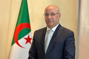 Tayeb Zitouni en costume avec drapeau algérien en arrière-plan.