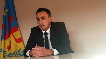 Aksel Bellabbaci en interview, portant un costume noir, avec drapeau kabyle en arrière-plan.