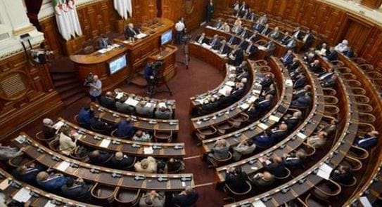Séance parlementaire avec des députés assis en cercle dans une salle en bois.