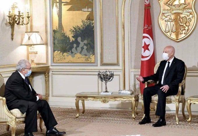 Deux responsables en réunion officielle, portant des masques, dans un décor élégant.