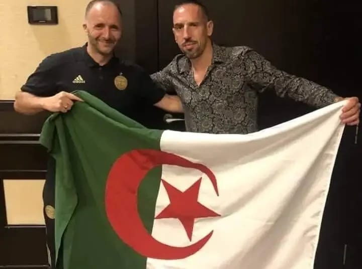 Deux hommes avec drapeau algérien, souriants, en intérieur.