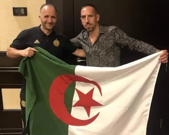 Deux hommes avec drapeau algérien, souriants, en intérieur.