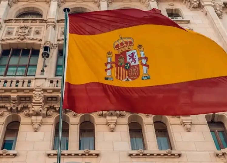 Nouveau dispositif de régularisation des sans-papiers en Espagne ! Drapeau espagnol flottant devant un bâtiment historique à Alger.