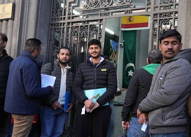 Groupe de migrants devant une ambassade avec drapeaux espagnol et pakistanais.