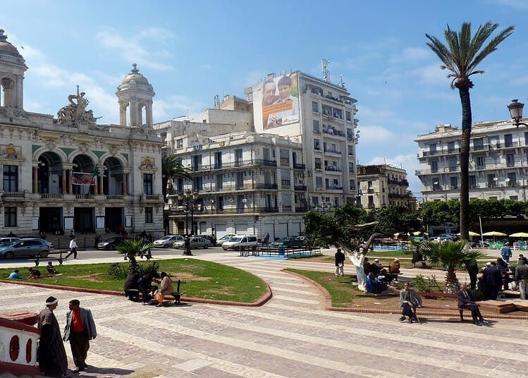 Vue d'une place animée avec bâtiments historiques et palmiers en Algérie.