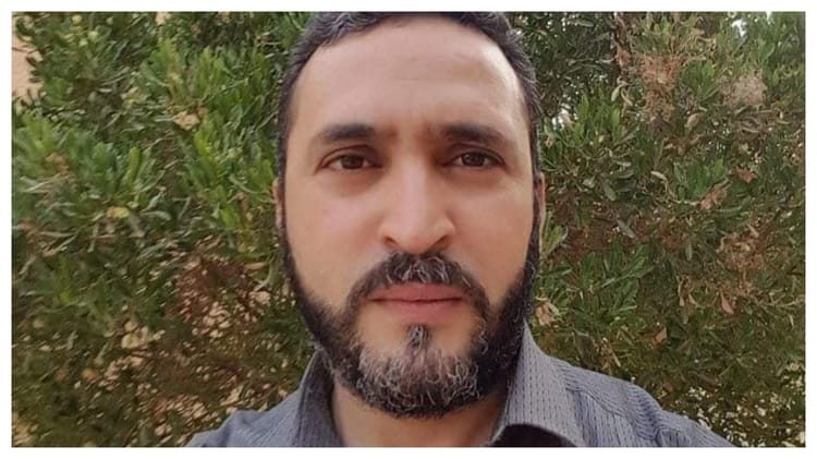 Homme avec barbe et cheveux courts, fond de feuillage vert.