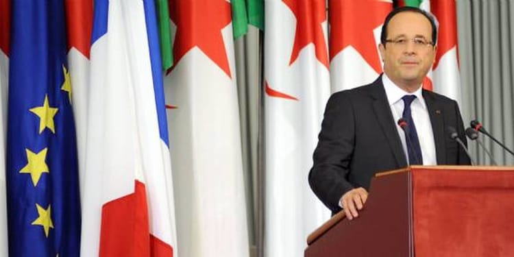 Homme politique parlant à une conférence avec drapeaux en arrière-plan.
