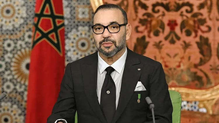 Homme en costume noir avec médaille, drapeau marocain en arrière-plan.