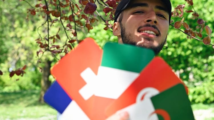 Cet algérien découvre une triste nouvelle après son trajet Paris – Alger à pied Jeune homme souriant sous un arbre avec cartes de voyage colorées.