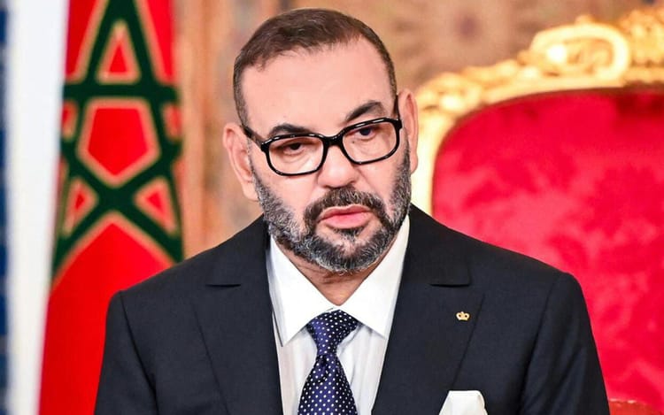 Homme en costume noir avec lunettes, siège dans un cadre officiel marocain.