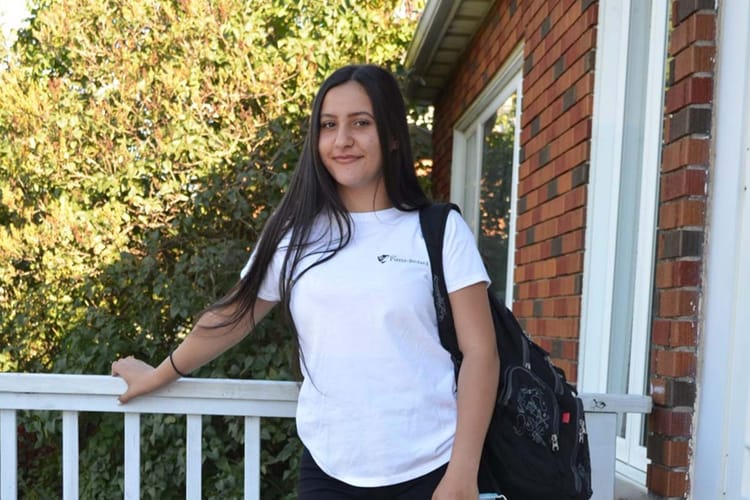 Jeune femme souriante avec sac à dos devant une maison en briques.