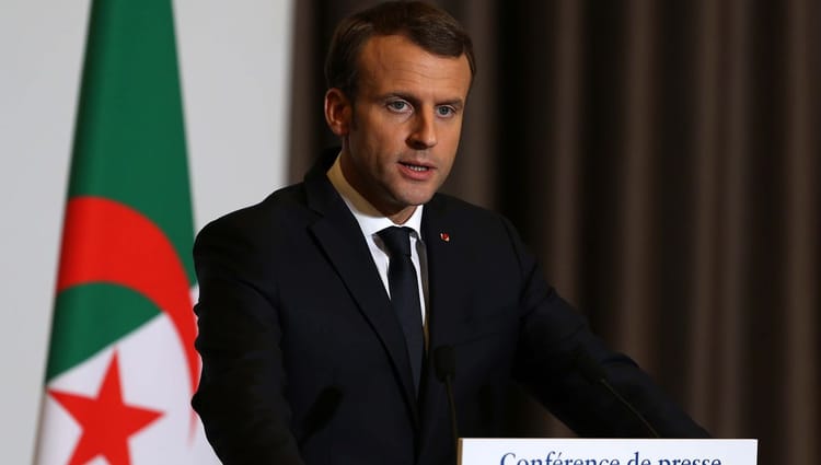 Emmanuel Macron parle lors d'une conférence avec drapeau algérien en arrière-plan.