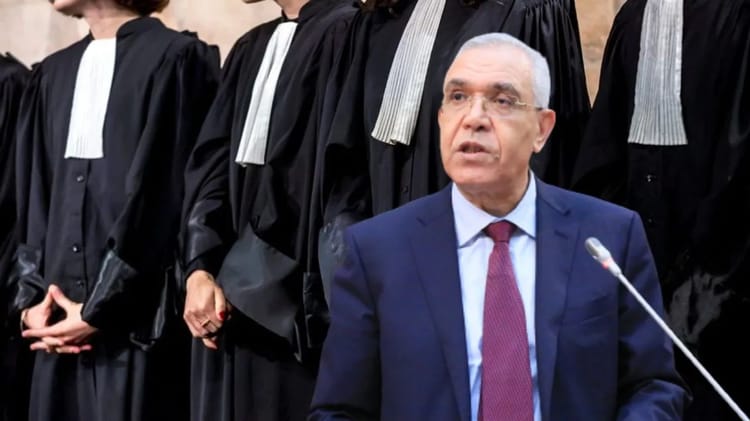 Homme en costume parlant lors d'une cérémonie avec des juges en robe noire.