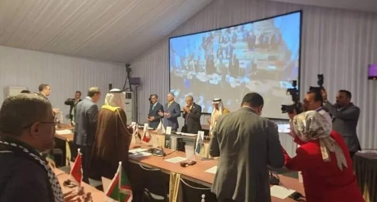 Réunion diplomatique avec discours en grand écran, participants en discussion.