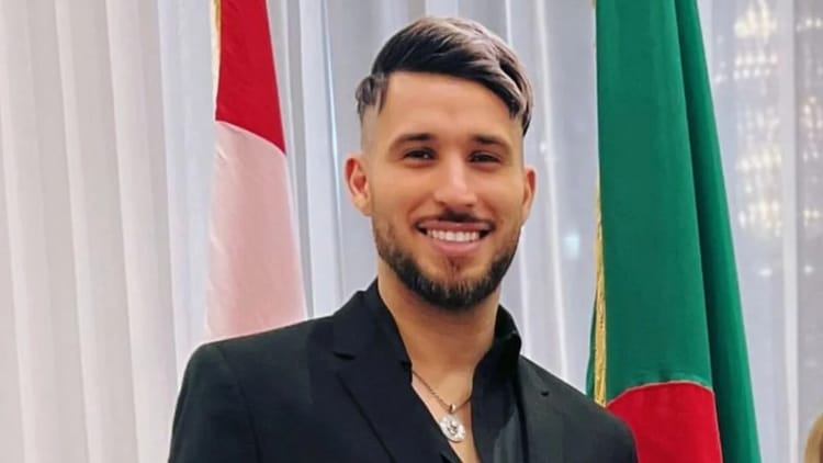 Fayçal El-Mignon souriant, portant un costume noir, devant un drapeau algérien et un drapeau étrangé.