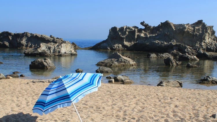 Touriste en Algérie : voici les codes à respecter pour éviter des problèmes Plage algérienne avec parasol bleu et rochers au bord de la mer.