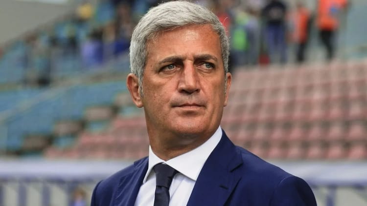 Homme en costume en stade, regard sérieux, contexte sportif.