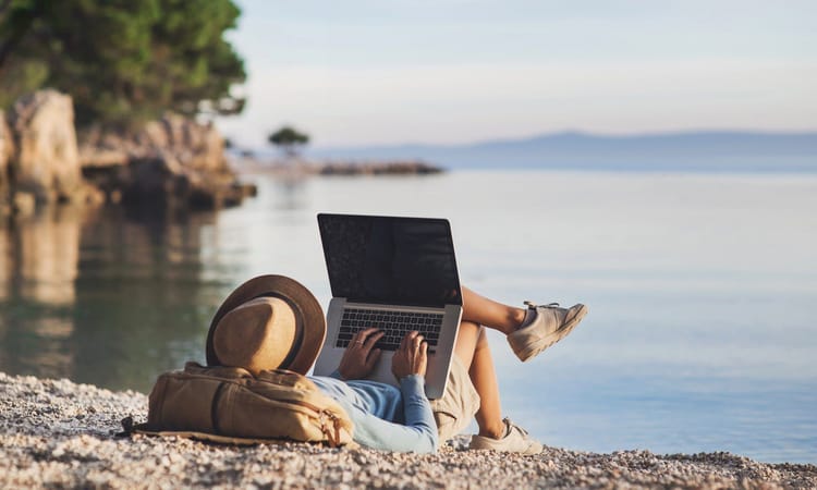 Tout ce que vous devez savoir sur le visa pour nomade numérique : avantages, pays participants et démarches Personne utilisant un ordinateur portable sur la plage avec un chapeau et un sac.
