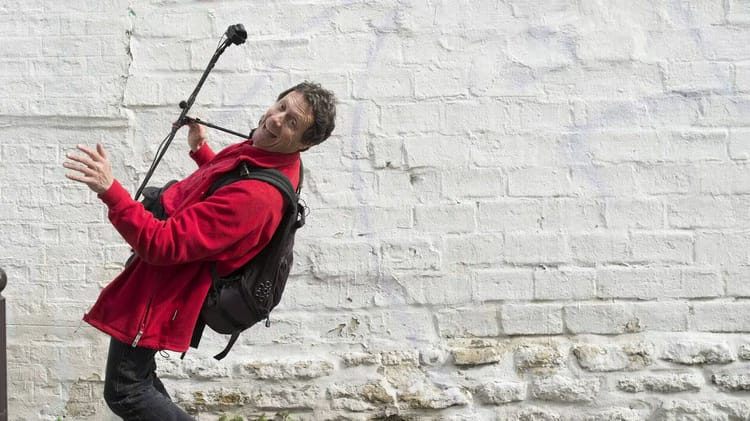 Découvrez l'aventure épique d'Antoine de Maximy en Algérie : un record historique pour "J'irai dormir chez vous" Antoine de Maximy avec un microphone dans un décor urbain blanc.