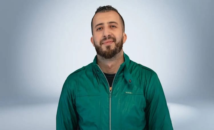 Anes Tina souriant, portant une veste verte, dans un studio neutre.