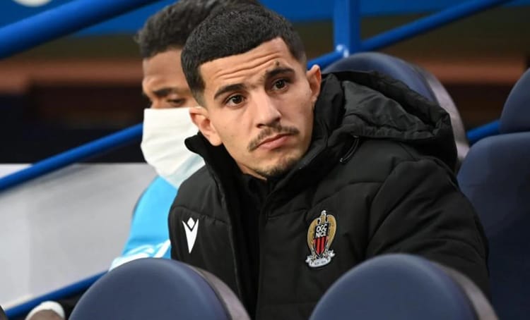 Youcef Atal assis sur le banc, portant un manteau noir avec le logo de Nice.