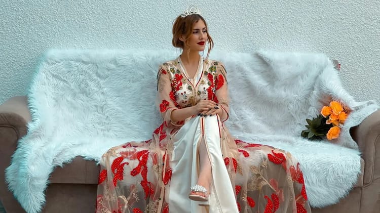 Femme portant un caftan marocain traditionnel avec broderies rouges et or, assise sur un canapé.
