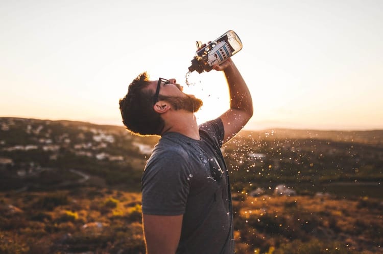 These Are The Best Watches From Around The Web This Week Homme portant des lunettes buvant une boisson en extérieur au coucher du soleil.
