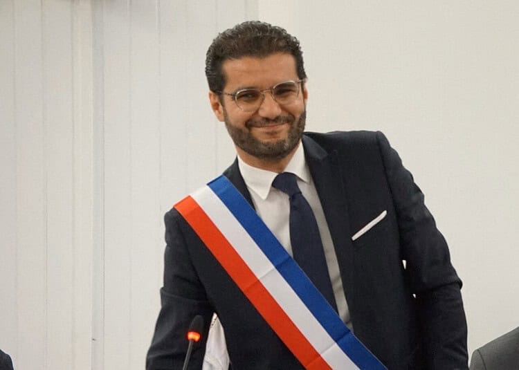 Un Franco-Algérien défie les attentes et devient maire ! Homme en costume avec écharpe tricolore, sourire confiant.