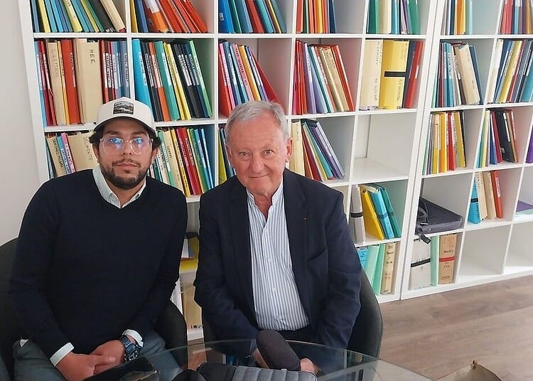 Expulsion controversée : un Algérien malade face à l'OQTF Deux hommes assis à une table en bibliothèque, livres en arrière-plan.